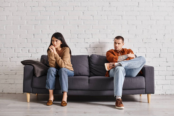 молодой человек смотрит на жену, сидящую на диване в гостиной, неприятности в отношениях - Фото, изображение