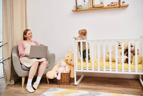 freudige Frau arbeitet am Laptop neben kleinem Sohn im Kinderbett im Kinderzimmer, Multitasking-Mutter - Foto, Bild
