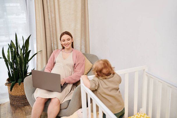 uśmiechnięta kobieta pracująca na laptopie w pobliżu małego synka w łóżeczku niemowlęcym w pokoju dziecinnym, wielozadaniowość - Zdjęcie, obraz
