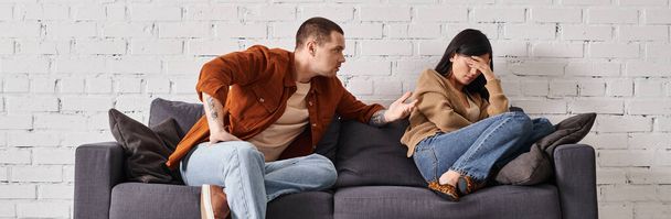 роздратований чоловік розмовляє з молодою азіатською дружиною плаче на дивані у вітальні, горизонтальний банер - Фото, зображення