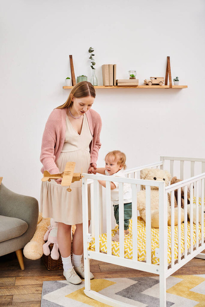 fröhliche schwangere Frau spielt mit Spielzeugflugzeug und Kleinkind im Kinderzimmer zu Hause, Mutterschaft - Foto, Bild