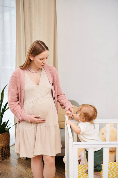 Küçük oğlunun elini tutan mutlu hamile kadın rahat bir bebek odasında beşikte duruyor, annelik. - Fotoğraf, Görsel