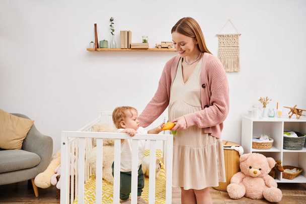 joyful pregnant woman holding toy while playing with little son standing in crib in nursery room - Photo, Image