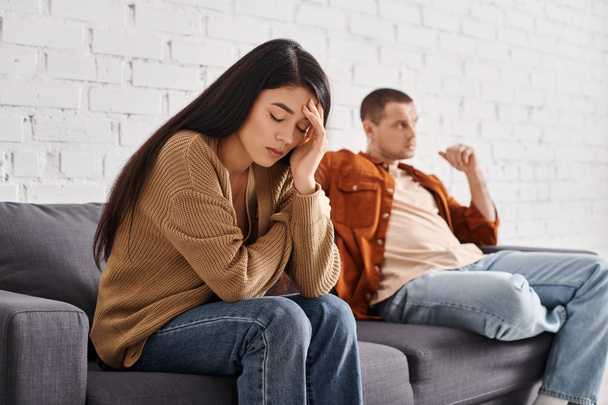 joven deprimido asiático mujer sentado con cerrado ojos cerca ofendido marido en sofá en sala de estar - Foto, Imagen