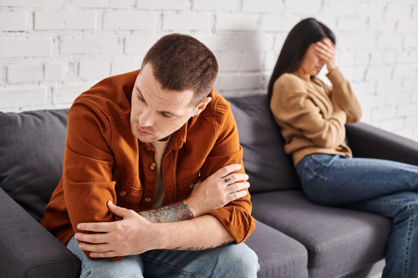 boos getatoeëerde man zit op de bank in de woonkamer in de buurt huilen aziatische vrouw, echtscheiding concept - Foto, afbeelding