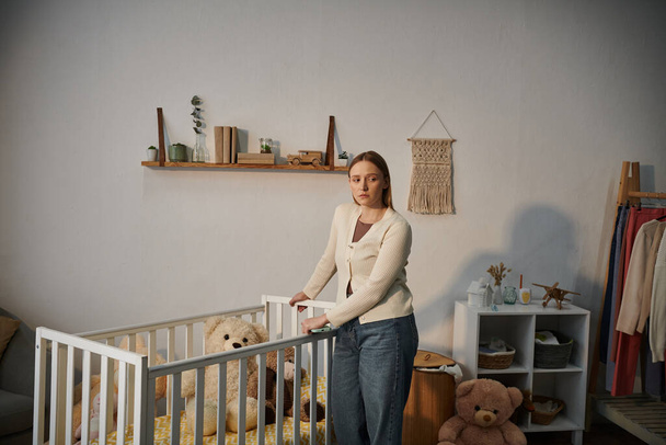 συγκλονισμένη και μοναχική νεαρή γυναίκα στέκεται κοντά κούνια με μαλακά παιχνίδια στο ζοφερό παιδικό δωμάτιο στο σπίτι - Φωτογραφία, εικόνα
