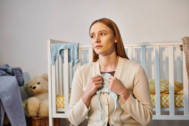 молода сумна жінка тримає дитячий одяг і дивиться далеко біля ліжечка в дитячій кімнаті вдома - Фото, зображення