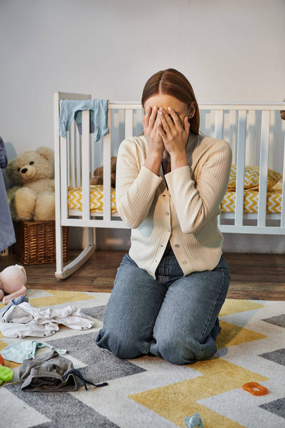 mladá a truchlící žena pláče v blízkosti dětské oblečení a postýlky s měkkými hračkami v dětském pokoji - Fotografie, Obrázek