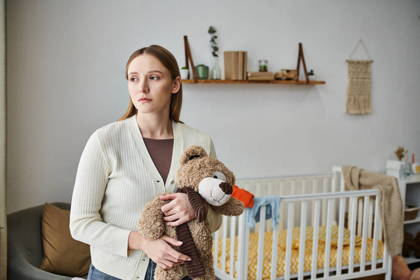 depressieve jonge vrouw met zacht speelgoed kijken weg in de kinderkamer thuis, gebroken hart moment - Foto, afbeelding
