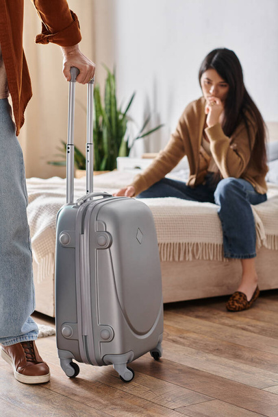 frustrovaná asijská žena sedí na posteli, zatímco její manžel s kufrem opouští domov, rozvod - Fotografie, Obrázek