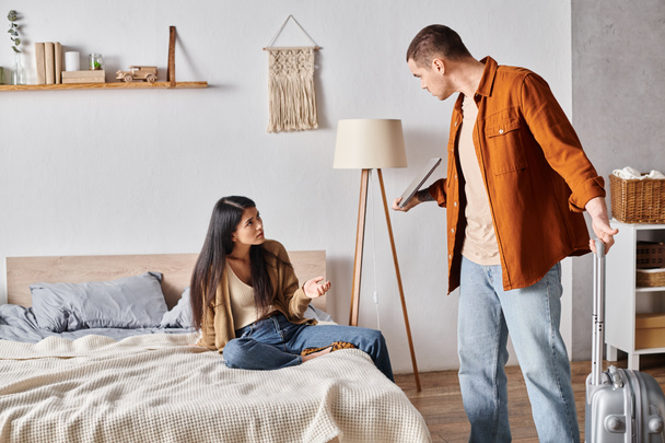 man with suitcase and laptop quarrelling with frustrated asian wife in bedroom, divorce concept - Photo, Image