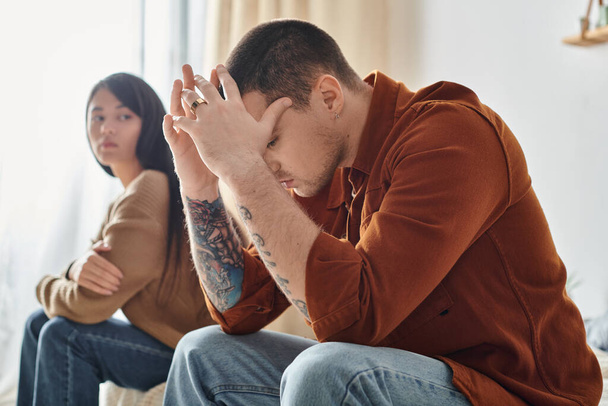 młody tatuaż zdenerwowany mężczyzna siedzi z łukiem głowa w pobliżu młody Azji kobieta, rodzina rozwód koncepcja - Zdjęcie, obraz