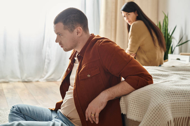 νεαρός απογοητευμένος άνθρωπος κάθεται στο πάτωμα κοντά αναστατωμένος Ασιάτισσα σύζυγος, δυσκολία στη σχέση έννοια - Φωτογραφία, εικόνα