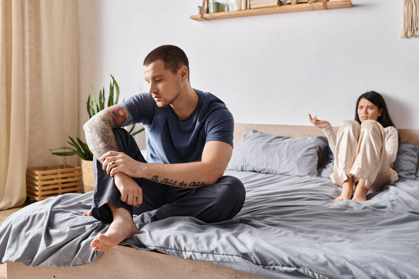 uražený tetovaný muž sedí na posteli v blízkosti hádky asijské manželky, rodinný vztah obtíže - Fotografie, Obrázek