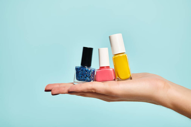 young unknown female model holding blue pink and yellow nail polish on her hand on blue backdrop - Photo, Image