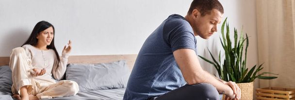 młody zdenerwowany wytatuowany mężczyzna siedzi na łóżku w domu w pobliżu kłótnia azjatyckie żona, poziome transparent - Zdjęcie, obraz