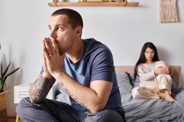mladý tetovaný muž dívá pryč, zatímco sedí na posteli v blízkosti uražené asijské manželky, rozvod koncept - Fotografie, Obrázek