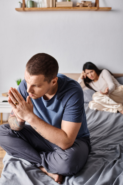 junge frustrierte tätowierte Mann sitzt auf dem Bett in der Nähe beleidigt asiatische Frau, Familie Scheidungskonzept - Foto, Bild