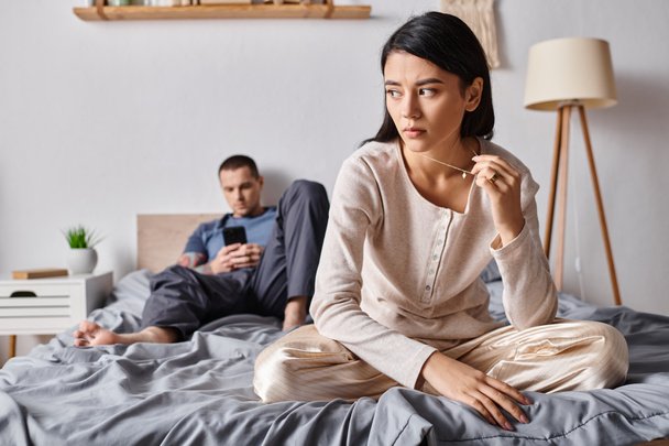 αναστατωμένος Ασιάτισσα γυναίκα κάθεται κοντά στο σύζυγο χρησιμοποιώντας smartphone στο υπνοδωμάτιο στο σπίτι, οικογενειακό πρόβλημα - Φωτογραφία, εικόνα