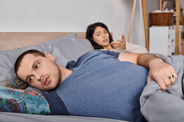 młody sfrustrowany wytatuowany mężczyzna odwracając wzrok leżąc na łóżku obok azjatyckiej żony, konflikt rodzinny - Zdjęcie, obraz