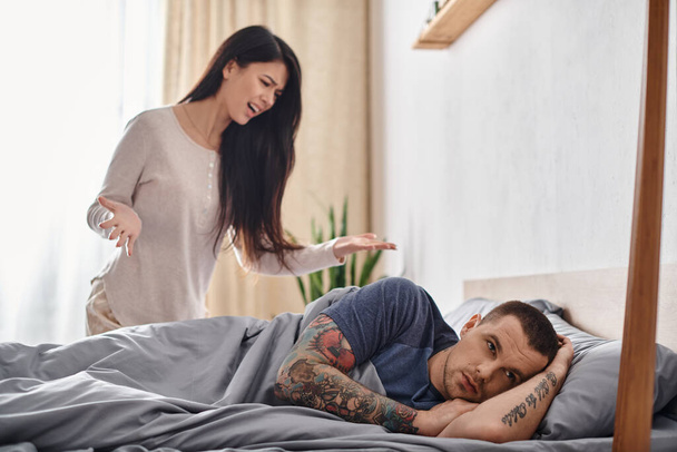 irritated asian woman quarreling at offended tattooed husband lying on bed at home, divorce concept - Photo, Image