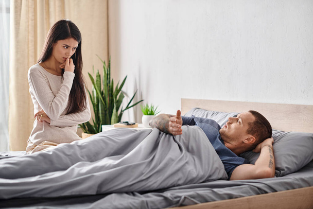obrażona Azji kobieta patrząc na młody wytatuowany mąż leżący na łóżku w domu, koncepcja rozwodu - Zdjęcie, obraz