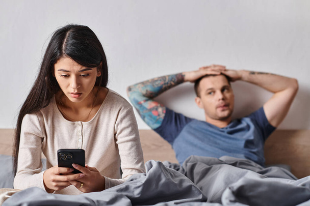 удивленный азиатская женщина, глядя на смартфон рядом муж лежит на кровати дома, семейный конфликт - Фото, изображение