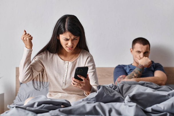 Wütende Asiatin schaut auf Smartphone neben Ehemann, der zu Hause im Bett liegt, Familienkonflikt - Foto, Bild