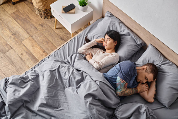 high angle view of asian wife and tattooed man lying on bed, relationship difficulties concept - Photo, Image