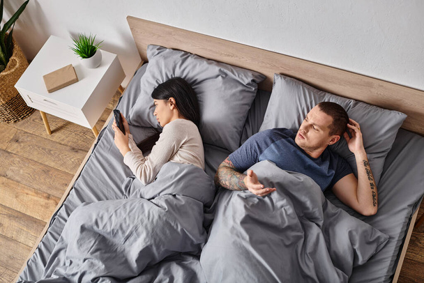 young asian woman looking in smartphone of husband lying on bed at home, family conflict concept - Photo, Image