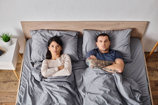 вид сверху на молодую разочарованную мультикультурную пару лежащую со сложенными руками на кровати дома - Фото, изображение