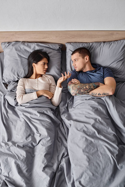 κορυφαία άποψη των νέων διαφυλετικό ζευγάρι ξαπλωμένος με διπλωμένα χέρια και φιλονικία στο κρεβάτι στο σπίτι - Φωτογραφία, εικόνα