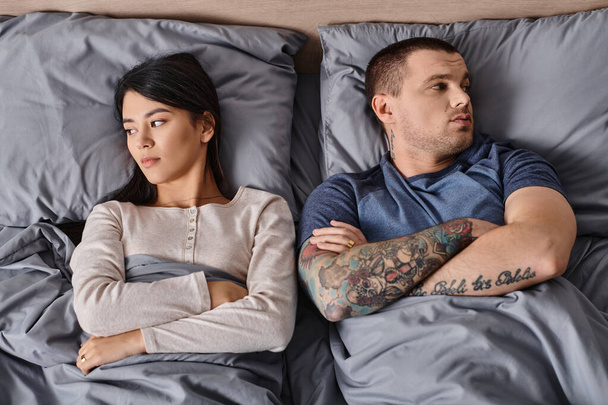 top view of depressed interracial couple lying down with folded arms and looking away on bed - Photo, Image