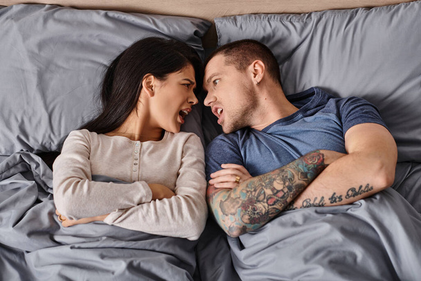 vista superior do casal interracial irritado deitado com os braços dobrados e brigando na cama em casa - Foto, Imagem