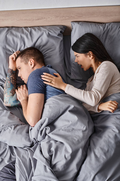ベッドに横たわるタトゥーの若い夫で口論している刺激的なアジアの女性のトップビュー, 離婚 - 写真・画像