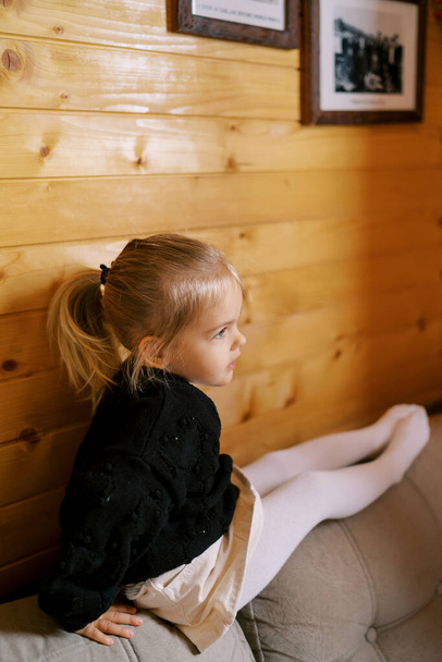 La niña se sienta en la parte posterior del sofá apoyada en una pared de madera y mira a la distancia. Foto de alta calidad - Foto, imagen