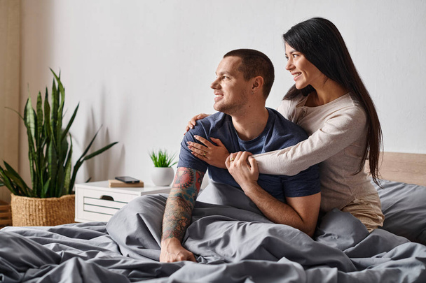 cheerful asian woman hugging young tattooed man looking away in bedroom at home, joy and happiness - Fotografie, Obrázek