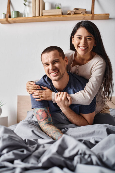 radostný multikulturní pár objímající a dívající se na kameru v ložnici doma, radost a štěstí - Fotografie, Obrázek