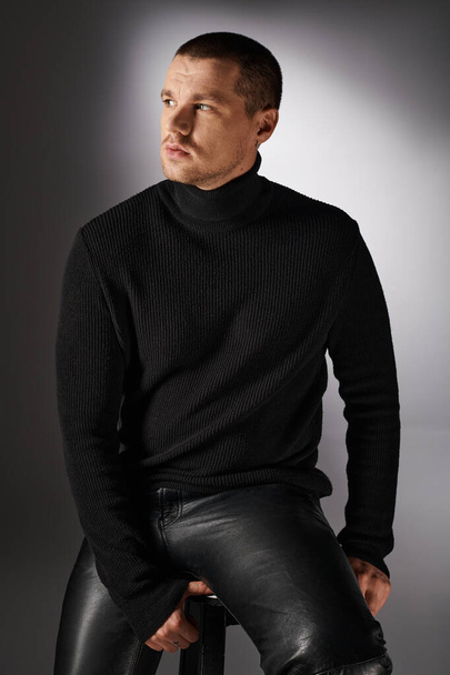 Goedkope trendy man in zwarte coltrui zittend en kijkend weg op grijze achtergrond met verlichting - Foto, afbeelding