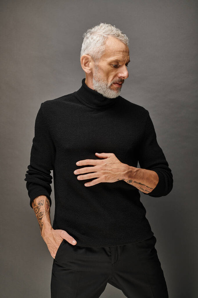 aantrekkelijke modieuze volwassen man in trendy coltrui poseren en weg te kijken op grijze achtergrond - Foto, afbeelding