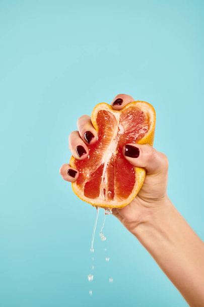 неизвестная молодая модель с лаком для ногтей, сжимающая свежий сочный грейпфрут на синем фоне - Фото, изображение