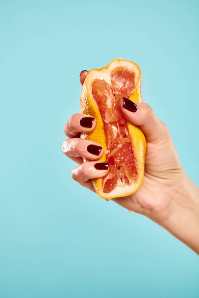 unknown woman with nail polish squeezing gourmet fresh grapefruit in her hand on blue background - Photo, Image