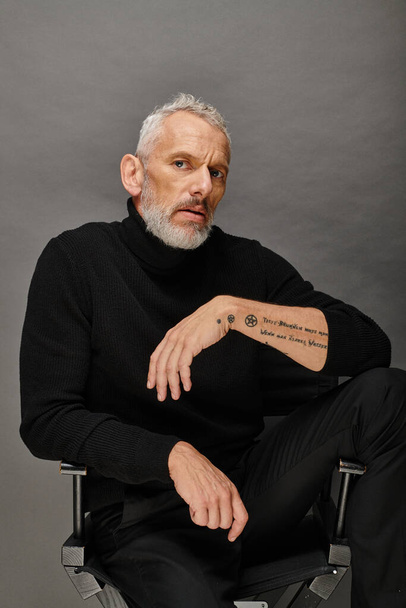 attrayant homme mature avec barbe à col roulé à la mode assis sur une chaise pliante et regardant la caméra - Photo, image