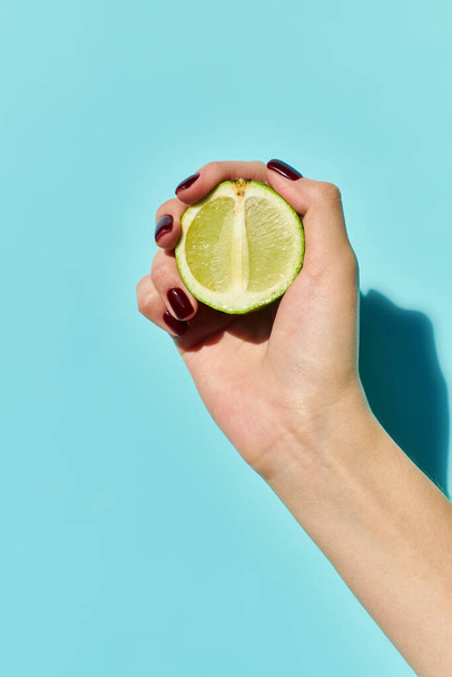 fresh green lime in hand of unknown young woman with nail polish posing on vibrant blue backdrop - Photo, Image