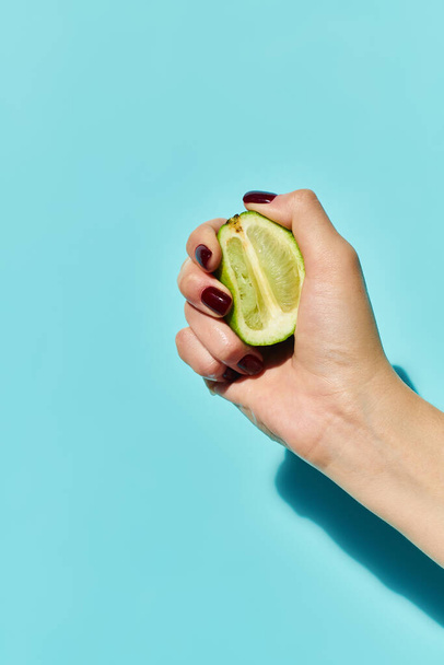 unknown female model with nail polish squeezing fresh ripe lime in hand on vibrant blue backdrop - Photo, Image