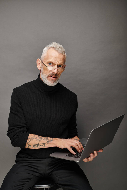 attractive mature man in trendy black turtleneck sitting on chair with laptop and looking at camera - Foto, Bild