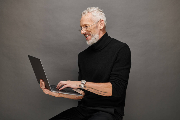 allegro bell'uomo maturo barbuto con gli occhiali seduto sulla sedia con computer portatile e sorridente felicemente - Foto, immagini