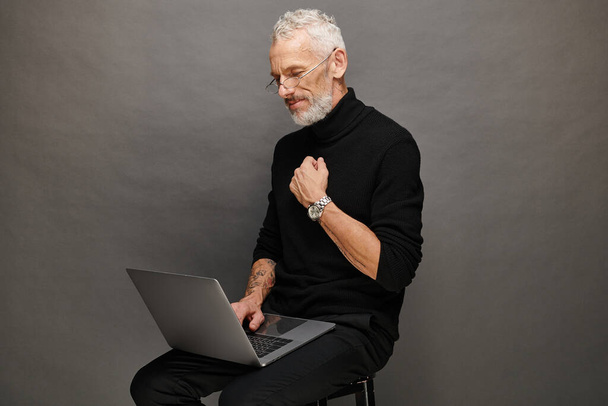 hombre maduro barbudo de buen aspecto en cuello alto negro sentado en la silla y mirando a su computadora portátil - Foto, imagen