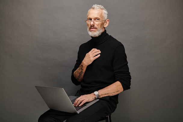 atractivo hombre maduro en la moda cuello alto negro sentado en la silla con el ordenador portátil y mirando a la cámara - Foto, imagen