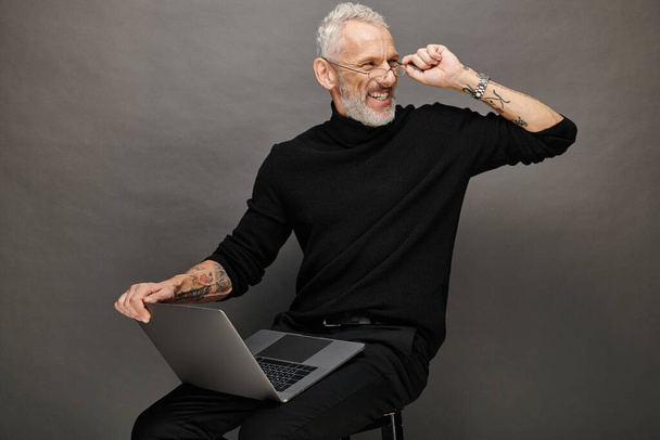 joyous attractive bearded mature man with glasses sitting on chair with laptop and smiling happily - Photo, Image